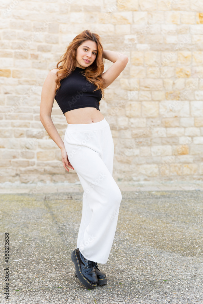 a wonderful pose of a beautiful young girl with long curly red hair wearing a black shirt and white pants and an ancient stone background