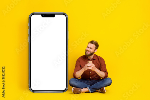 Full size portrait of cheerful man sit floor hold telephone look empty space banner isolated on yellow color background