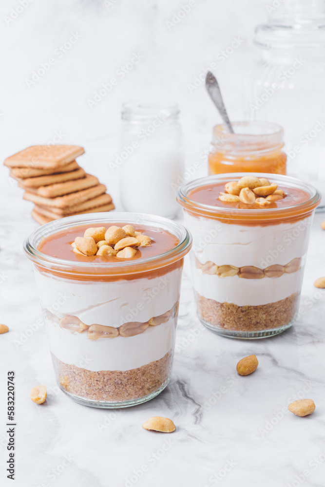 Cheesecake in a jar. Layers of grinded cookies, caramel, peanuts and cream cheese. Covered in caramel and decorated with peanuts. Selective focus. Macro shot. Cheesecake in a jar. Layers of grinded cookies, caramel, peanuts and cream cheese. Covered in caramel and decorated with peanuts. Selective focus. Macro shot.