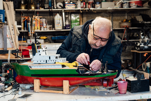 Retired elderly man making ship model building handcraft on the table in his garage. Pensioner assembling boat model with screwdriver. Active Retirement lifestyle, hobby. Selective focus.