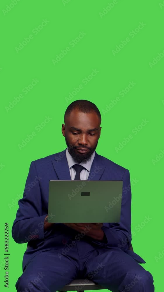 Vertical video: Front view of african american manager working on laptop in studio, sitting on chair with wireless pc. Corporate employee in office business suit using portable computer over green Vertical video: Front view of african american manager working on laptop in studio, sitting on chair with wireless pc. Corporate employee in office business suit using portable computer over green