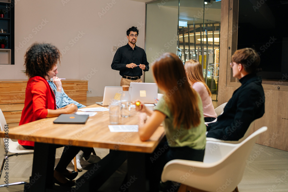 Wide shot of professional male speaker giving corporate presentation for diverse businesspeople at meeting room, presents new business plan. Startup team sitting at boardroom at office listening boss.