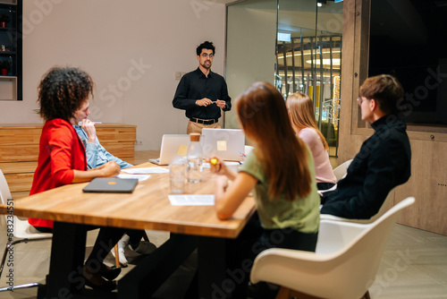 Wide shot of professional male speaker giving corporate presentation for diverse businesspeople at meeting room, presents new business plan. Startup team sitting at boardroom at office listening boss.