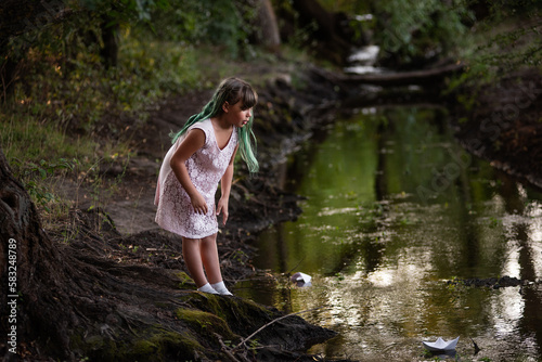 Teenage girl with green hair in dress, stands on the river bank, launches white paper origami boat