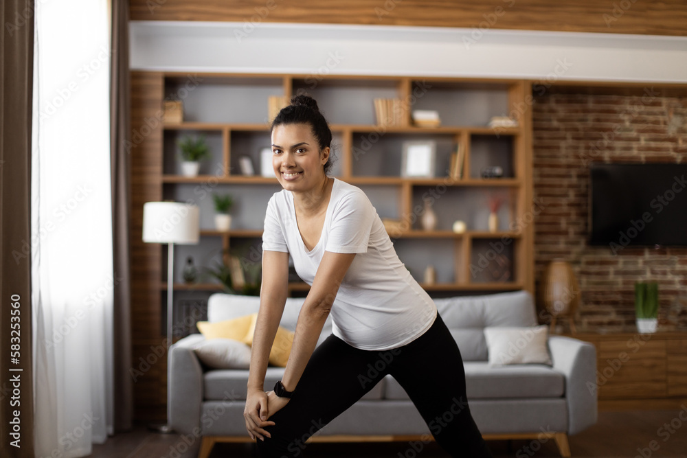 Fototapeta premium African american woman with dark hair stretching legs during workout at modern living room. Sporty female in activewear smiling and looking at camera while training during free time.