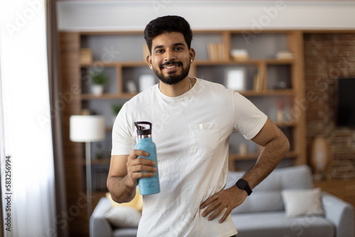 Sporty indian man smiling sincerely on camera while standing in living room with sport bottle of water in hand. Handsome guy wearing active clothes during exercises.