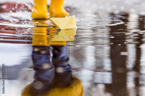 It's springtime. A small child in yellow rubber boots jumps through puddles, plays and launches paper boats on the water.. Photo of spring and autumn holidays. The concept of spring.