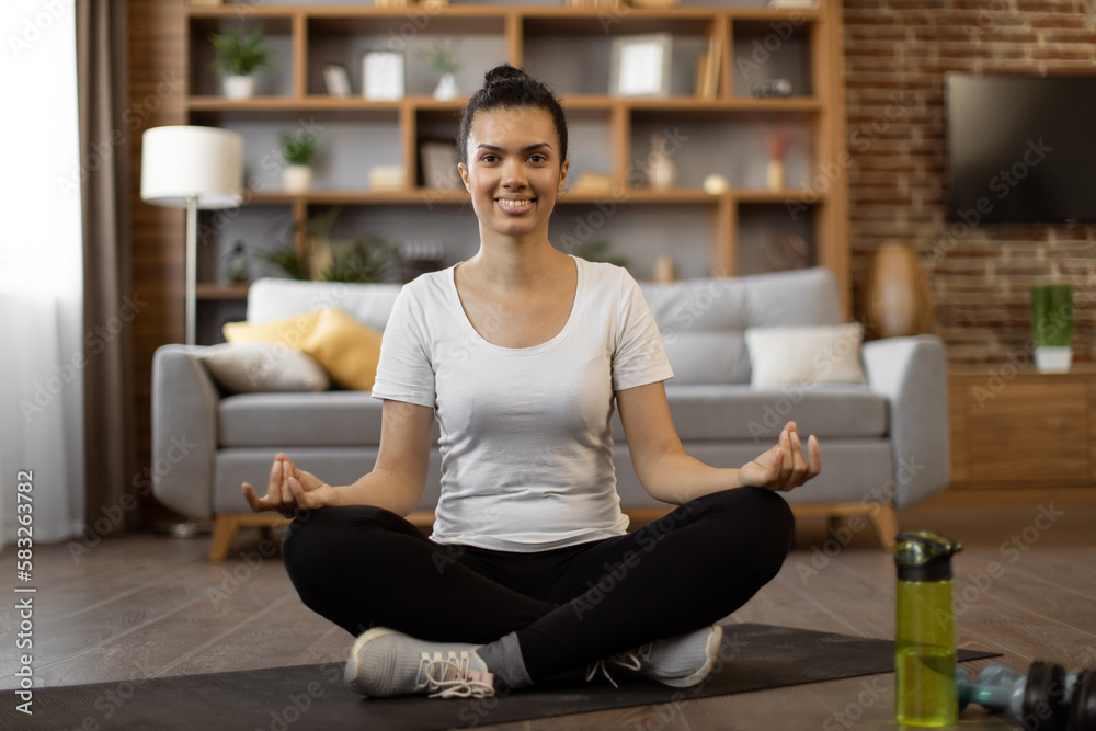 Portrait of african young woman in activewear sitting in lotus position with mudra gesture and smiling at camera. Happy beautiful female meditating at cozy apartment during free time.