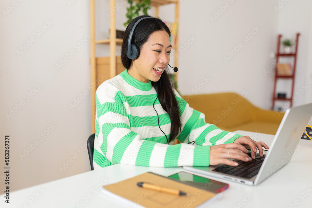 Young asian woman using headset looking at laptop screen while learning online at home. Cheerful chinese female with headphones having a video call for customer call center service Young asian woman using headset looking at laptop screen while learning online at home. Cheerful chinese female with headphones having a video call for customer call center service