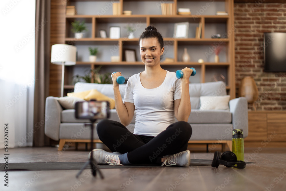 Smiling multiracial fitness trainer recording tutorial of weight lifting with dumbbells above head using camera of smartphone on tripod indoors. Active trainer filming video content for sports vlog. Smiling multiracial fitness trainer recording tutorial of weight lifting with dumbbells above head using camera of smartphone on tripod indoors. Active trainer filming video content for sports vlog.