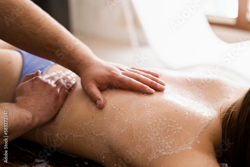 Salt scrub massage on back of girl in spa salon close-up. Masseur making cosmetic scrubbing and peeling procedure for skin rejuvenation.