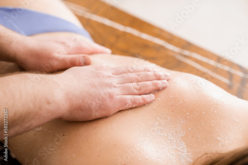 Masseur making cosmetic scrubbing and peeling procedure for skin rejuvenation. Salt scrub massage on back of girl in spa salon close-up.