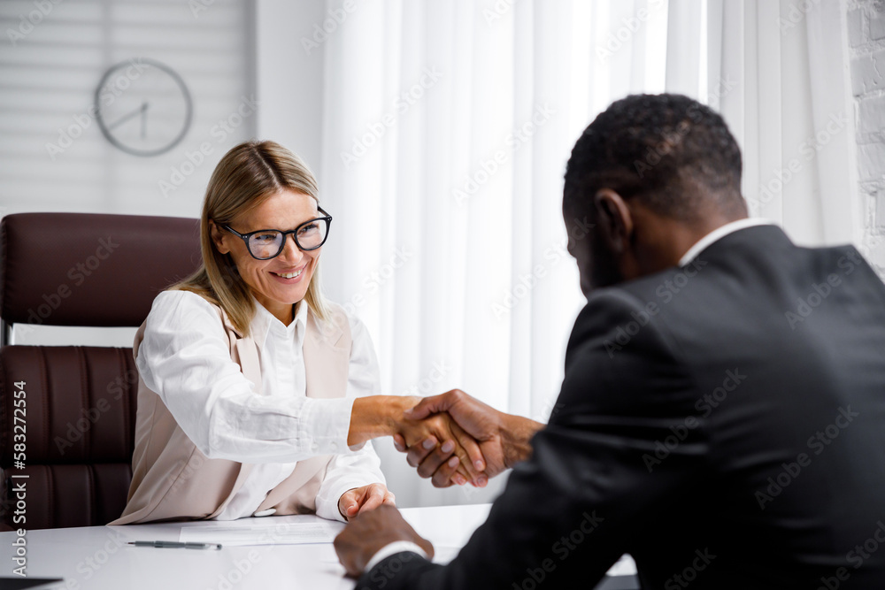 Hiring, interview. Middle-aged blond business woman conducting an interview in a bright office, an African American man. He looks at his resume and after reading it, shakes hands and smiles. Hiring, interview. Middle-aged blond business woman conducting an interview in a bright office, an African American man. He looks at his resume and after reading it, shakes hands and smiles.