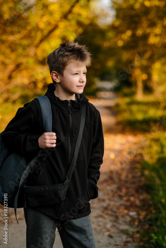Vertical portrait of European fair-haired a teenager in black jacket with backpack on background of orange trees, looking straight, happy young man in park, place for text, autumn photo, side view
