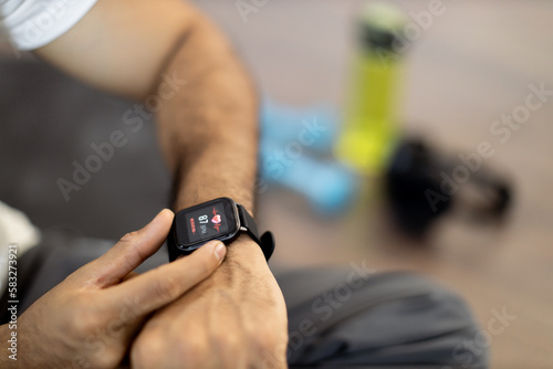 Close-up of young sporty indian man sitting on wooden floor and checking pulse on modern smartwatch with sports app after workout at home. Athletic male leading healthy lifestyle by training indoors.