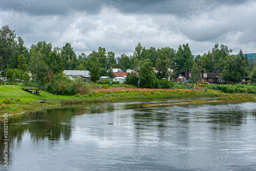 Fairbanks, Alaska, USA - July 27, 2011: Chena River shoreline opposite Griffin Park under gray cloudscape. Green belt separates water from sky. Red roofs peeks through green foliage © Klodien