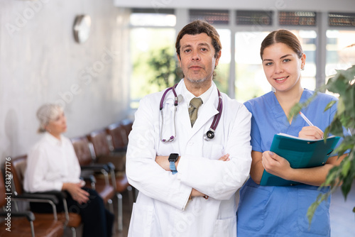 Confident experienced doctor in white coat standing with young female assistant in modern medical office