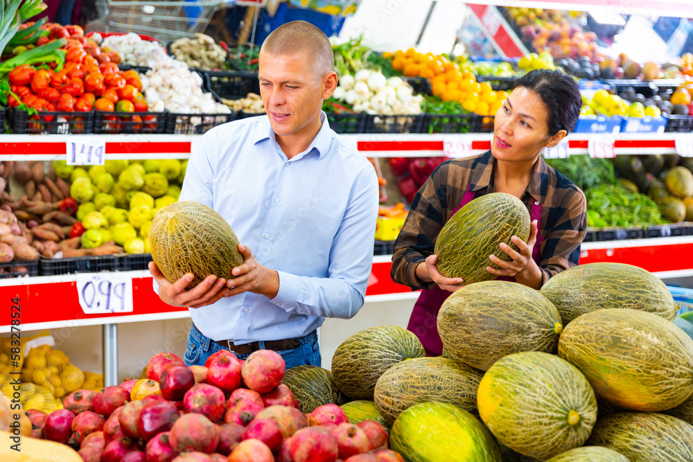 Friendly asian saleswoman helping male buyer to choose ripe sweet melon in fruit and vegetable store..