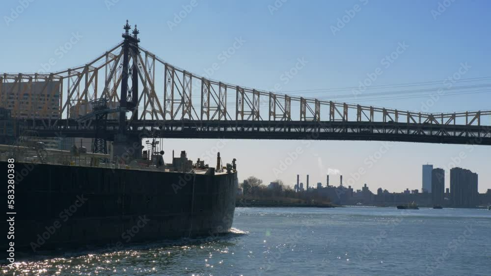 New York, Roosevelt Island Bridge. A large long barge floats down the river under a bridge over which cars drive, with New York skyscrapers in the background. New York, America - March 02, 2020. New York, Roosevelt Island Bridge. A large long barge floats down the river under a bridge over which cars drive, with New York skyscrapers in the background. New York, America - March 02, 2020.