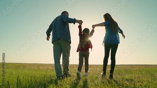 Happy family is playing in park at sun, child holding hands is jumping. Teamwork. Dad mom child play together in meadow. Active child daughter, family playing in park. Little girl running with parents