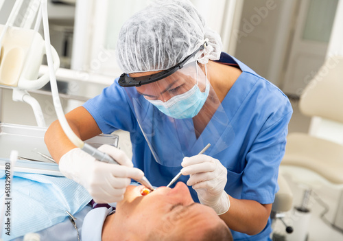 Dentist in mask and face shield treating male patient using dental drill in dentist office