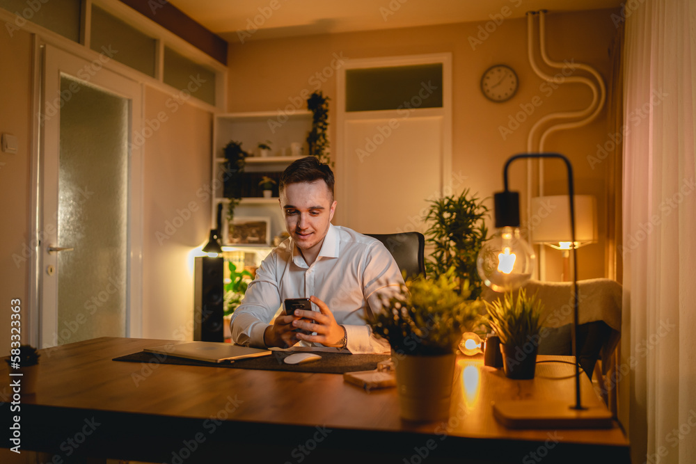 A young man is working late or studying at his home office while drinking coffee and using his mobile phone A young man is working late or studying at his home office while drinking coffee and using his mobile phone