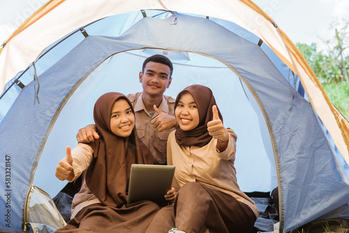boy scout and two girl scouts in veil using tablets with thumbs up in a tent on nature