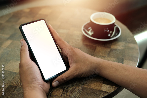 Mockup image of a man holding mobile phone with blank white screen with coffee cup on wooden table