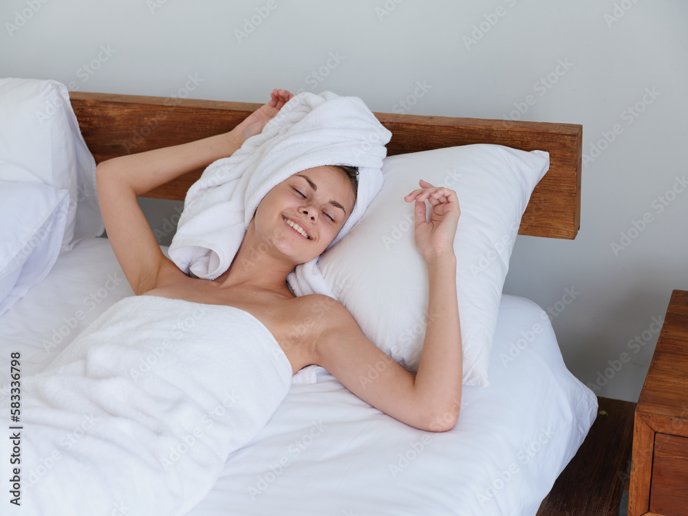 A woman lies on the bed after a shower with wet hair with a towel on her head in the bedroom and strokes her body skin, a cute smile with teeth and weekend rest in bed, women's beauty day, lifestyle A woman lies on the bed after a shower with wet hair with a towel on her head in the bedroom and strokes her body skin, a cute smile with teeth and weekend rest in bed, women's beauty day, lifestyle