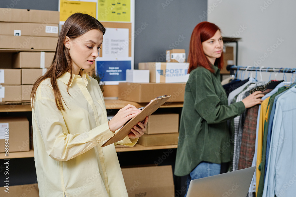 Obraz premium Young brunette woman with document looking through list of people needing new warm clothes against her colleague standing by rack
