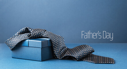 Fathers Day text background banner. blue gift box with ribbon bow and necktie on dark blue table. Monochrome. Gift delivery for him. Male fashion