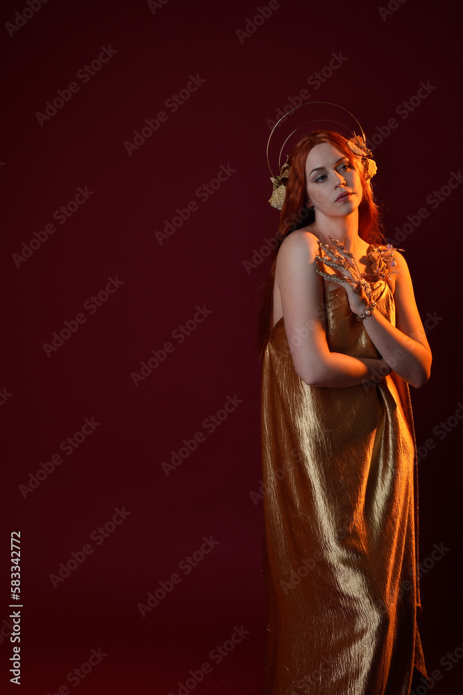 Close up fantasy portrait of beautiful woman model with red hair, goddess silk robes & gold crown.  Posing with gestural hands reaching out, isolated on dark red studio background 