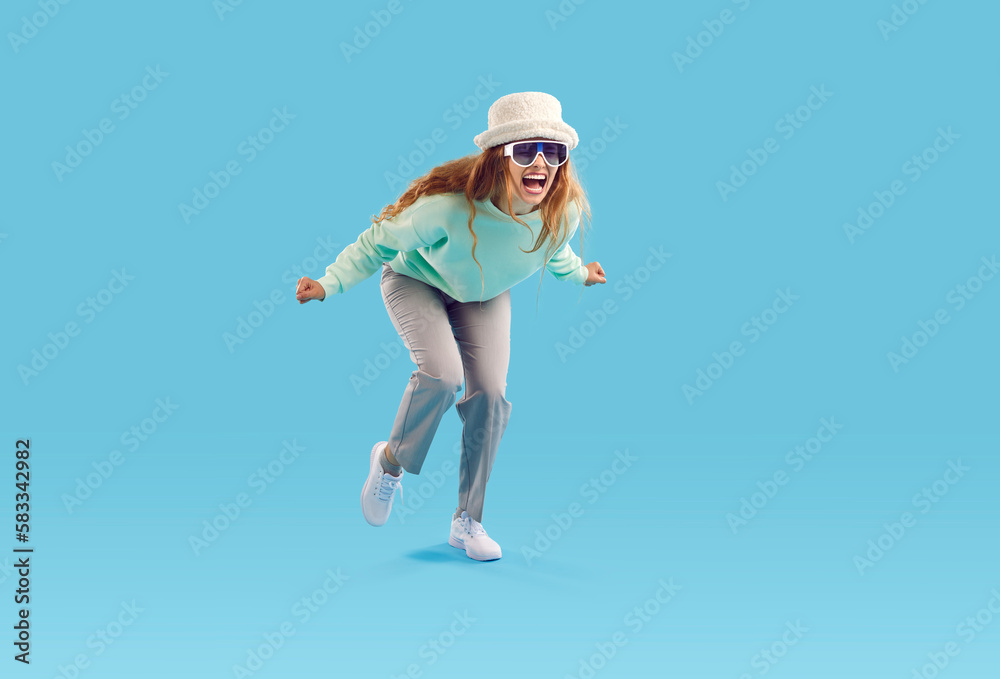 Studio shot of funny crazy screaming girl in fashionable outfit. Young woman in hat and sunglasses feeling happy and excited or furious and enraged screaming loudly isolated on blue background