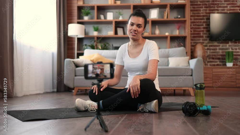 Fit african american sports blogger conducting remote fitness exercises while sitting on floor in front of digital device on tripod. Healthy young woman encouraging extra physical training at home. Fit african american sports blogger conducting remote fitness exercises while sitting on floor in front of digital device on tripod. Healthy young woman encouraging extra physical training at home.