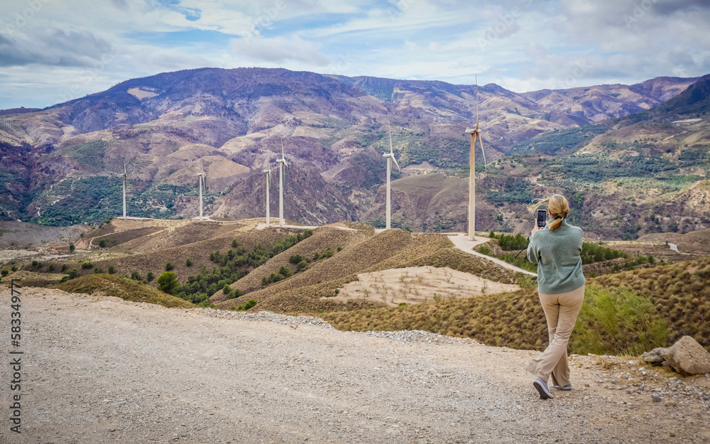 Blond young woman taking photos of a mountain landscape with wind turbines