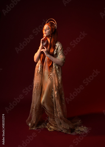 Full length fantasy portrait of beautiful woman model with red hair  goddess silk robes   gold crown. Standing pose gestural hands reaching out isolated on dark red studio background 