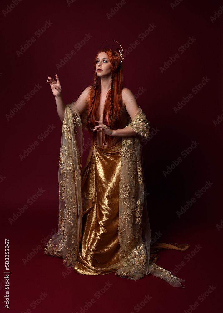 Full length fantasy portrait of beautiful woman model with red hair, goddess silk robes & gold crown. Standing pose gestural hands reaching out isolated on dark red studio background 