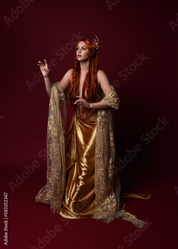 Full length fantasy portrait of beautiful woman model with red hair, goddess silk robes & gold crown. Standing pose gestural hands reaching out isolated on dark red studio background 