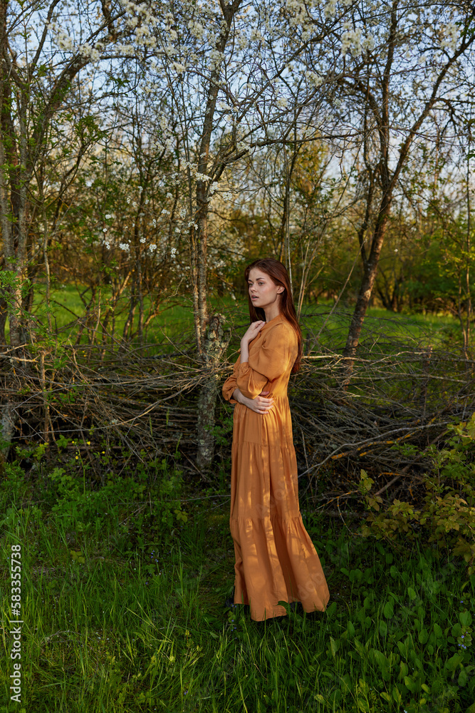 an elegant, sophisticated woman poses relaxed standing near a wicker fence at the dacha in a long orange dress enjoying the silence and peace