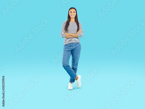 Smiling brunette young woman standing with folded hands. Studio portrait of happy stylish girl wearing casual jeans and t-shirt with long sleeves posing over light blue background