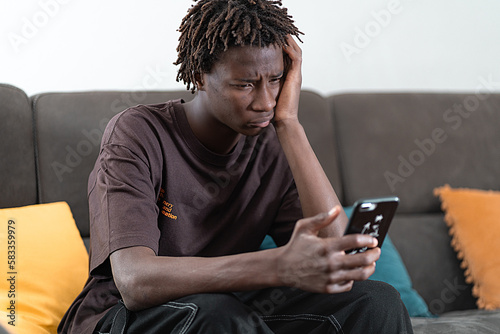 giovane ragazzo afroamericano seduto sul divano con lo smartphone in mano, fa un esprezione triste. photo