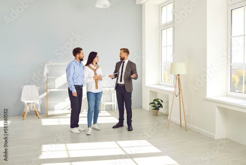 Handsome bearded man of a realtor in a suit with documents in his hands advises a young couple before buying a new house. Three people standing in a modern sunny apartment.