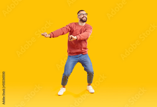 Cheerful, playful, funny and positive bearded man in glasses, red sweatshirt and jeans in good mood having fun dancing on yellow background, full length portrait. Banner for advertisement, marketing.