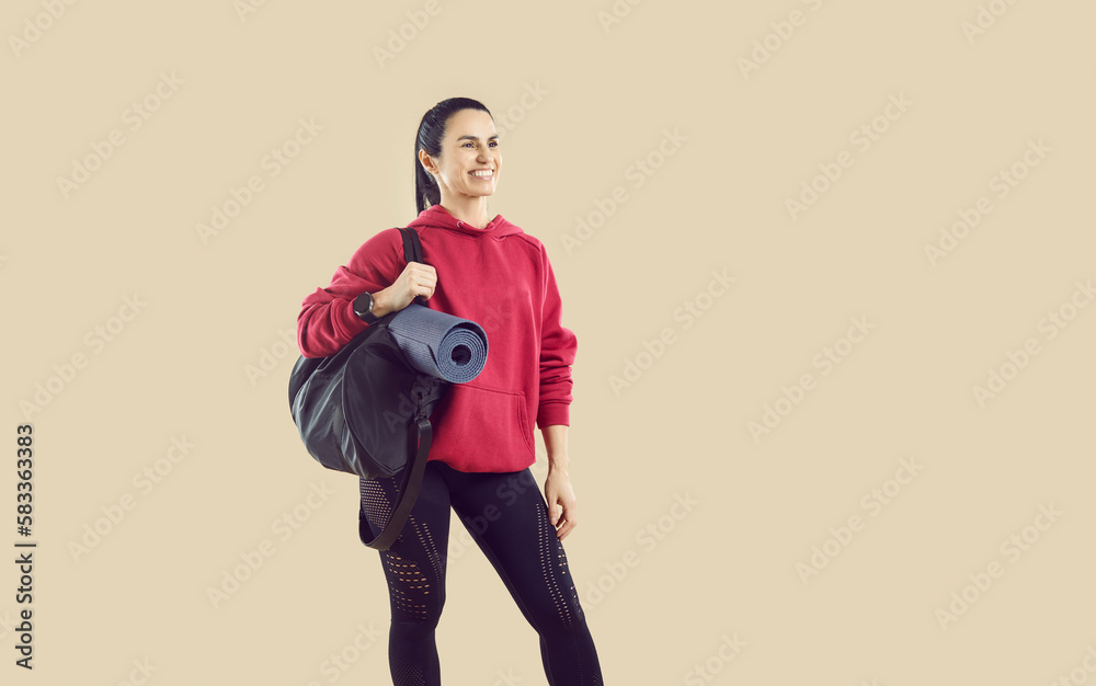 Fototapeta premium Portrait of happy smiling female athlete holding sports bag and workout mat. Beautiful fit woman wearing comfortable red hoodie and black leggings standing isolated on light beige copyspace background