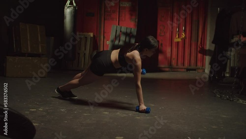 Determined athletic pretty fitness Asian female practicing weight training, exercising dumbbell renegade row, developing upper body strength while working out in garage gym.