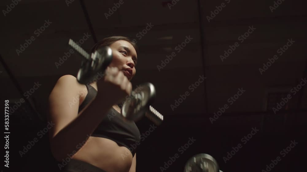 Portrait of determined motivated sporty fit attractive Asian woman practicing weight training , performing dumbbell bicep curls workout while exercising in dark gym. Low angle view.