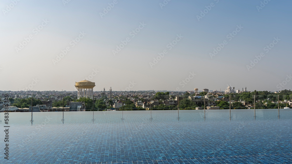 Infinity outdoor swimming pool on the roof. The tiled blue bottom is visible through the water. A transparent barrier along the edge. In the distance - a panorama of the city against the sky. India