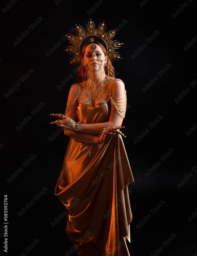 Close up fantasy portrait of beautiful woman model with red hair, goddess silk robes & ornate gold crown.  Posing with gestural hands reaching out, isolated on dark  studio background 