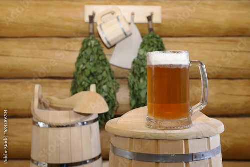 In the bath house, on an upside-down wooden barrel, there is a glass mug of light beer against the background of a log wall with traditional sauna accessories. 