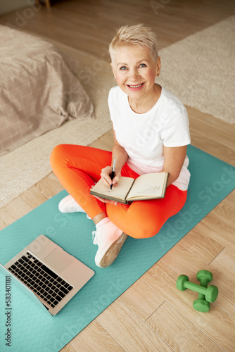High angle view of aged female in sports clothes looking at camera with smile, studying online, watching fitness tutorial on laptop sitting on blue mat in bedroom, noting down information in note pad